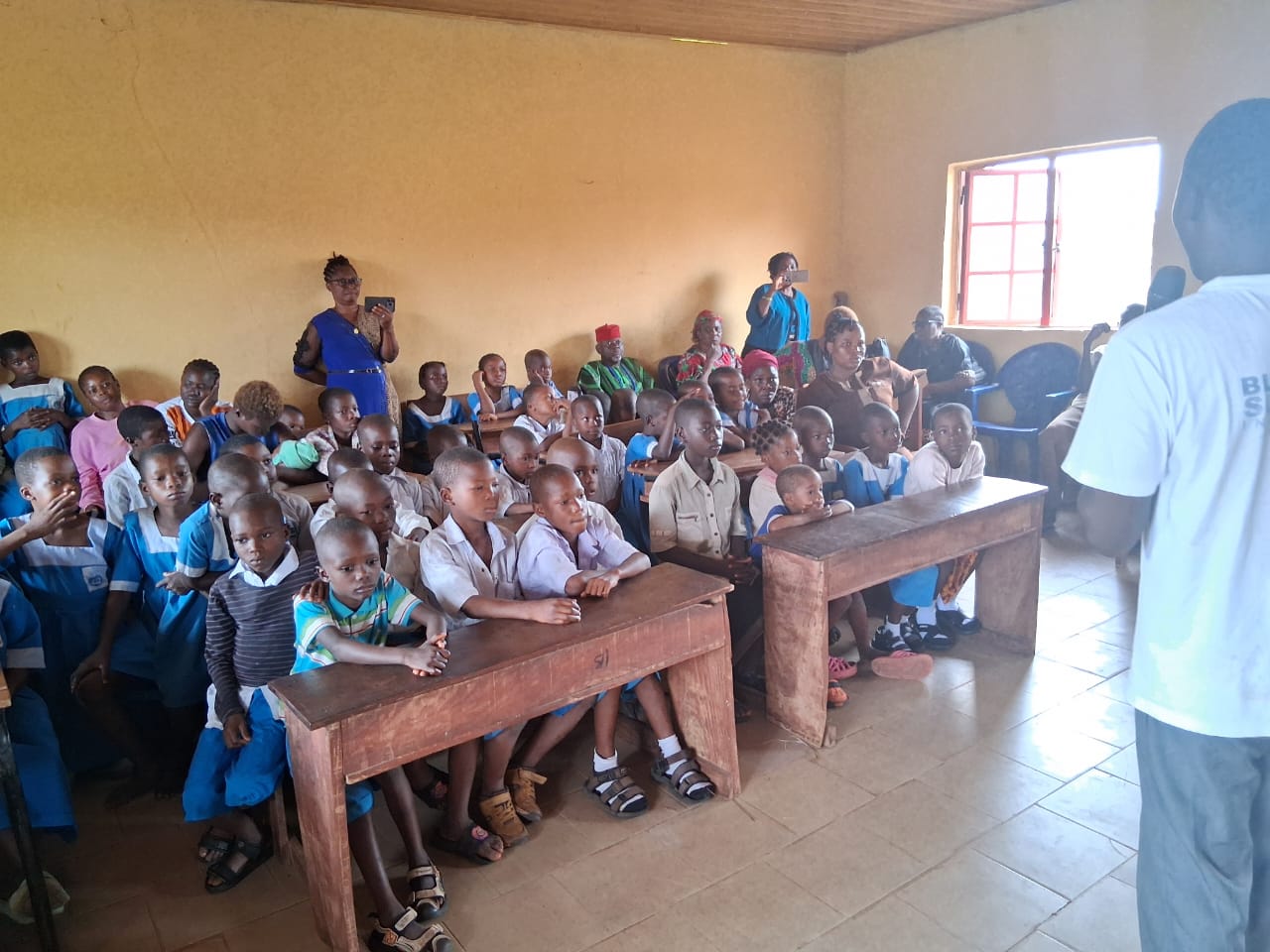 Pupils and community members seated at wooden desks in a classroom during the outreach